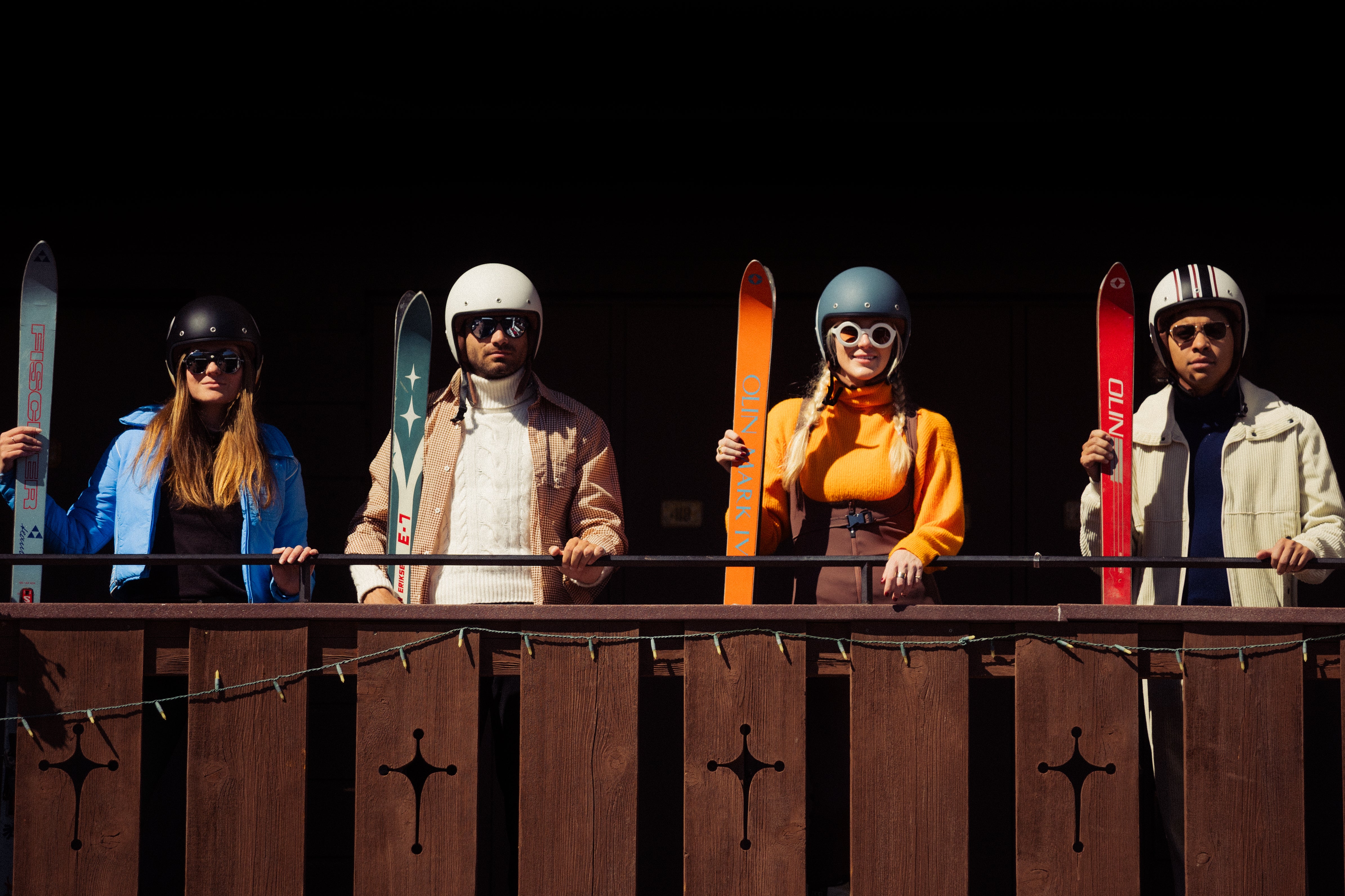 Four people with skis and helmets standing behind a wooden railing.